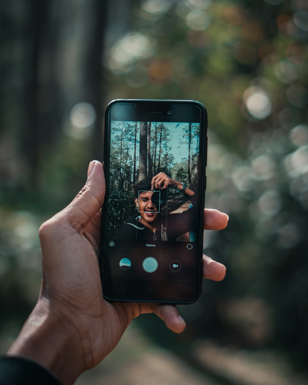 Persona sosteniendo un teléfono inteligente negro mientras toma una selfie debajo del árbol