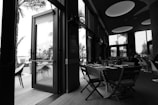 A black and white photograph of a modern indoor dining area with a table set for a meal. The open door reveals an outdoor seating area with chairs and palm trees, providing a view of a distant city skyline.