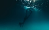 A person is diving underwater, surrounded by a dark, deep blue environment. Light filters down from the surface, creating a shimmering effect in the water above.