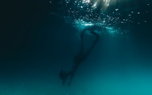 A person is diving underwater, surrounded by a dark, deep blue environment. Light filters down from the surface, creating a shimmering effect in the water above.