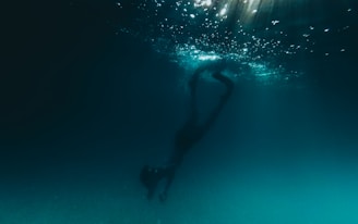 A person is diving underwater, surrounded by a dark, deep blue environment. Light filters down from the surface, creating a shimmering effect in the water above.