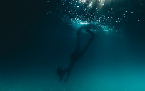 A person is diving underwater, surrounded by a dark, deep blue environment. Light filters down from the surface, creating a shimmering effect in the water above.