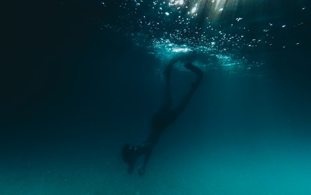A person is diving underwater, surrounded by a dark, deep blue environment. Light filters down from the surface, creating a shimmering effect in the water above.