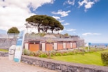 A beautifully landscaped area featuring a modern public facility with natural stone walls, surrounded by well-maintained greenery and large, leafy trees under a bright blue sky. A signboard stands near a paved path, and the ocean is faintly visible in the background.