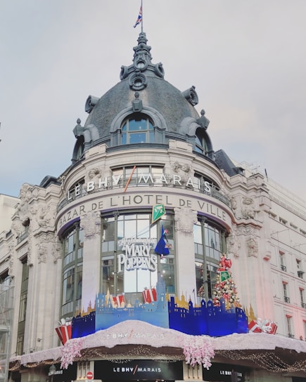 The image features a grand building with ornate architectural details, including intricate stonework and a large dome. The facade is adorned with festive decorations, including a Christmas tree and large presents. Signage for LE BHV MARAIS and BAZAR DE L'HÔTEL DE VILLE is prominently displayed. A banner advertising a Mary Poppins event is visible.