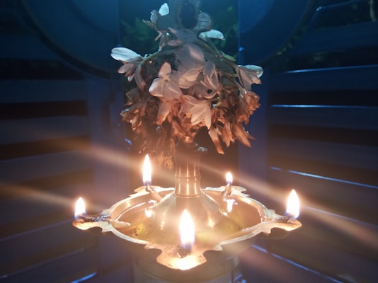 A brass oil lamp with five lit flames is adorned with jasmine flowers on top. The soft glow of the lamps creates a warm and serene ambiance, lighting up the dark surroundings with a peaceful aura. The lamp is placed against a backdrop of wooden panels, adding to the traditional aesthetic.