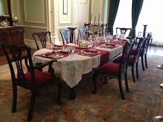 An elegant dining room set with a long wooden table, covered with a lace tablecloth and red placemats. There are multiple chairs around the table, all with red upholstered seats. The table is decorated with silver domes, glassware, and candles. The room features ornate wall moldings and intricate carpet with floral patterns. Large windows with green curtains provide natural light.