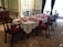 An elegant dining room set with a long wooden table, covered with a lace tablecloth and red placemats. There are multiple chairs around the table, all with red upholstered seats. The table is decorated with silver domes, glassware, and candles. The room features ornate wall moldings and intricate carpet with floral patterns. Large windows with green curtains provide natural light.