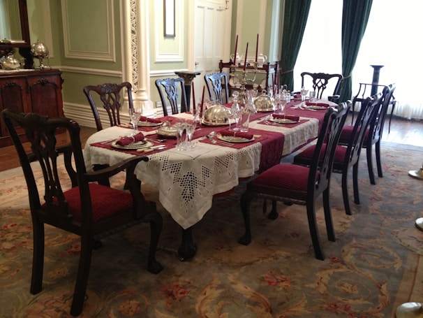 An elegant dining room set with a long wooden table, covered with a lace tablecloth and red placemats. There are multiple chairs around the table, all with red upholstered seats. The table is decorated with silver domes, glassware, and candles. The room features ornate wall moldings and intricate carpet with floral patterns. Large windows with green curtains provide natural light.
