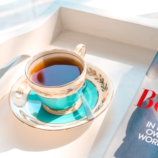 A decorative teacup filled with tea rests on a matching saucer with a floral design, placed on a white tray. A small silver spoon is beside the cup. Part of a magazine with bold red lettering is visible next to the tray.
