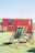 A bright red shipping container with a chalkboard sign on the side stands on a green artificial grass surface. In front of it, several wooden deck chairs with striped fabric are arranged casually. The sky is clear and blue, and some urban structures are visible in the background.