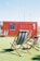 A bright red shipping container with a chalkboard sign on the side stands on a green artificial grass surface. In front of it, several wooden deck chairs with striped fabric are arranged casually. The sky is clear and blue, and some urban structures are visible in the background.
