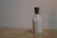 A small glass bottle with a cork stopper contains a fine white granular substance, possibly sand or salt. The bottle is placed on a plain wooden surface against a neutral wall background, and the lighting is soft and diffused.