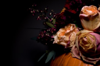 A close-up of a floral arrangement featuring dried roses with faded pink and orange petals. Darker flowers and green leaves are visible in the background, creating a contrast with the predominantly dark background.