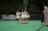 A group of female lacrosse players wearing white uniforms with red numbers and protective eyewear, embracing in celebration on a green field. A lacrosse goal with an orange frame is visible to the right, and the surrounding area is lined with trees.