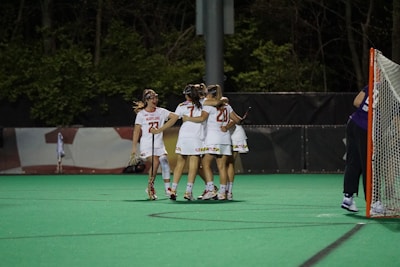 A group of female lacrosse players wearing white uniforms with red numbers and protective eyewear, embracing in celebration on a green field. A lacrosse goal with an orange frame is visible to the right, and the surrounding area is lined with trees.