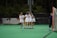 A group of female lacrosse players wearing white uniforms with red numbers and protective eyewear, embracing in celebration on a green field. A lacrosse goal with an orange frame is visible to the right, and the surrounding area is lined with trees.