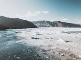 An expansive arctic landscape with a large glacier in the midground, surrounded by rugged mountains in the distance under a bright blue sky. The foreground features icy waters with chunks of ice scattered across the surface.