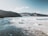 An expansive arctic landscape with a large glacier in the midground, surrounded by rugged mountains in the distance under a bright blue sky. The foreground features icy waters with chunks of ice scattered across the surface.