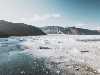 An expansive arctic landscape with a large glacier in the midground, surrounded by rugged mountains in the distance under a bright blue sky. The foreground features icy waters with chunks of ice scattered across the surface.