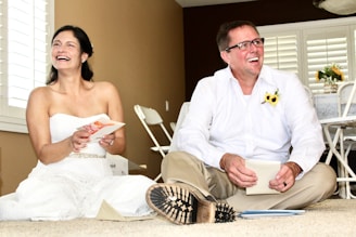 A woman in a white dress and a man in a white shirt and beige pants are sitting on the floor, smiling and laughing. The woman holds a greeting card, and the man has a sunflower pinned to his shirt. Behind them are white shutters on the windows and some foldable chairs against a brown wall. There is a table with a tablecloth and a vase with flowers in the background.