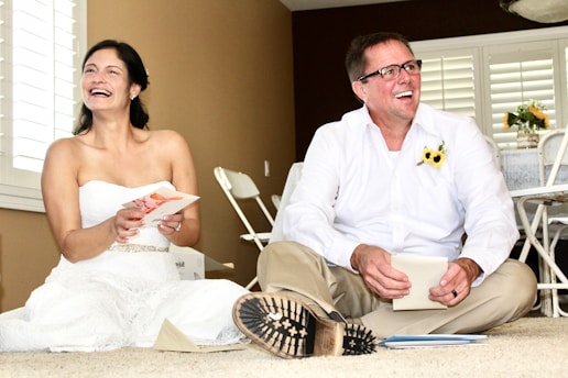 A woman in a white dress and a man in a white shirt and beige pants are sitting on the floor, smiling and laughing. The woman holds a greeting card, and the man has a sunflower pinned to his shirt. Behind them are white shutters on the windows and some foldable chairs against a brown wall. There is a table with a tablecloth and a vase with flowers in the background.
