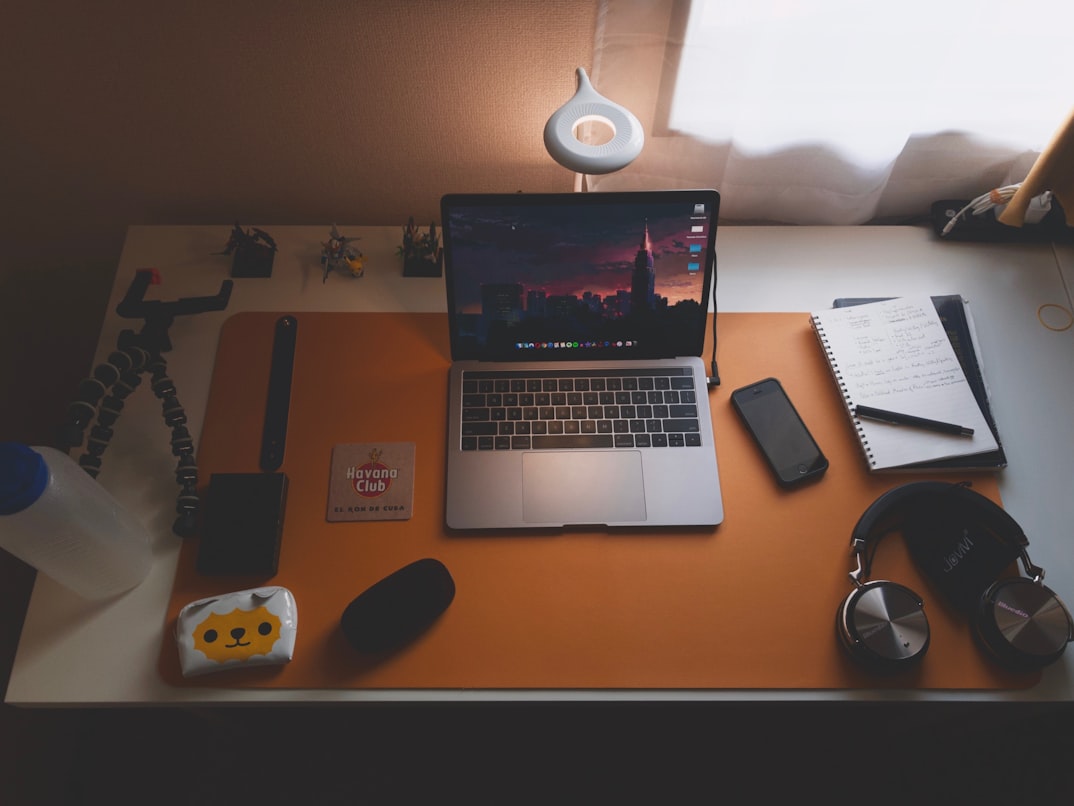 macbook pro with displaying a purple/orange sunset filled with mountains, standing among a cellphone, headphone, tripod and other work related stuff on a white table