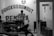 A black and white photograph of a barbershop with a person visible through the window. The shop seems small and cluttered with minimal furniture, and a motorcycle is partially visible in the foreground. The door has a sign that reads 'Buka,' indicating that the shop is open.