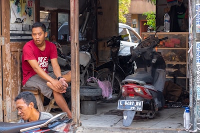 A man in a red t-shirt sits on a wooden stool at the entrance of a workshop or garage. Another man is working on a motorcycle near the foreground. Inside the garage, there are various tools and motorcycles, including a couple of mopeds. The workshop looks somewhat cluttered, with parts and equipment scattered around.