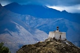 A solitary white stupa adorned with prayer flags is perched atop a rocky hill. The backdrop features towering, rugged mountains shrouded in clouds, creating a serene and dramatic landscape.