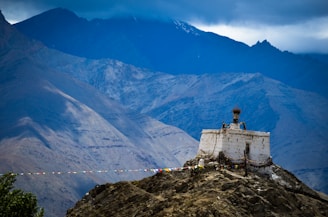 A solitary white stupa adorned with prayer flags is perched atop a rocky hill. The backdrop features towering, rugged mountains shrouded in clouds, creating a serene and dramatic landscape.