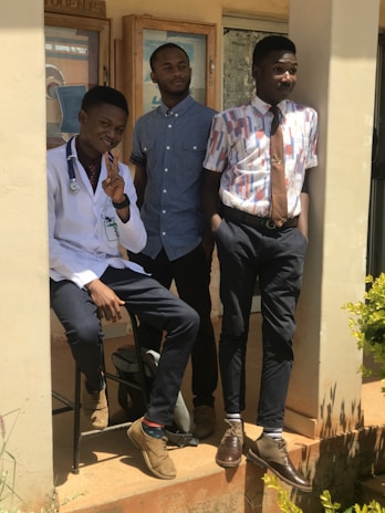 Three young men are posing together outside a building. One is wearing a white coat with a stethoscope, suggesting he might be a medical professional. The other two are dressed in casual, yet stylish attire, with button-up shirts and ties. They are standing on a porch with plants nearby, in bright sunlight.