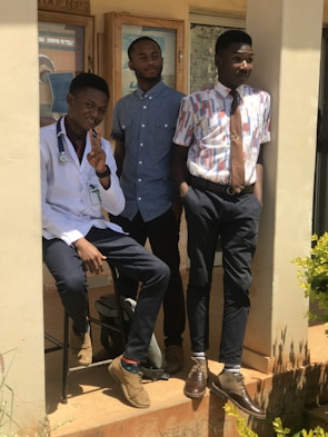 Three young men are posing together outside a building. One is wearing a white coat with a stethoscope, suggesting he might be a medical professional. The other two are dressed in casual, yet stylish attire, with button-up shirts and ties. They are standing on a porch with plants nearby, in bright sunlight.