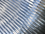Reflective metallic panels are arranged diagonally, capturing the image of the sky with scattered clouds. The panels have a sleek, modern appearance with perforated textures near the edge.