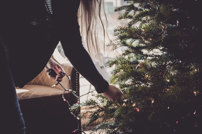 A person decorates an evergreen tree with a string of lights. Soft light filters through a nearby window, illuminating the room. An upholstered chair with a patterned fabric is positioned to the left.