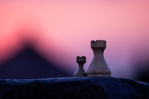 Two chess rooks are placed on a rock with a blurred background in shades of pink and purple, evoking a serene and contemplative atmosphere.
