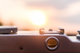 Close-up view of a vintage camera's top components with a clear focus on the adjustment dials and hot shoe. The background features a bright, glowing sunset or sunrise that softly blurs into an orange or pink gradient.