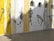 A mural featuring abstract figures painted in monochromatic tones. The figures are in various dynamic poses along a wall segmented by vertical lines in yellow and white. Sunlight casts shadows across the mural, creating an interplay of light and shadow.