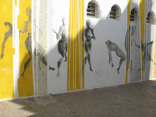 A mural featuring abstract figures painted in monochromatic tones. The figures are in various dynamic poses along a wall segmented by vertical lines in yellow and white. Sunlight casts shadows across the mural, creating an interplay of light and shadow.