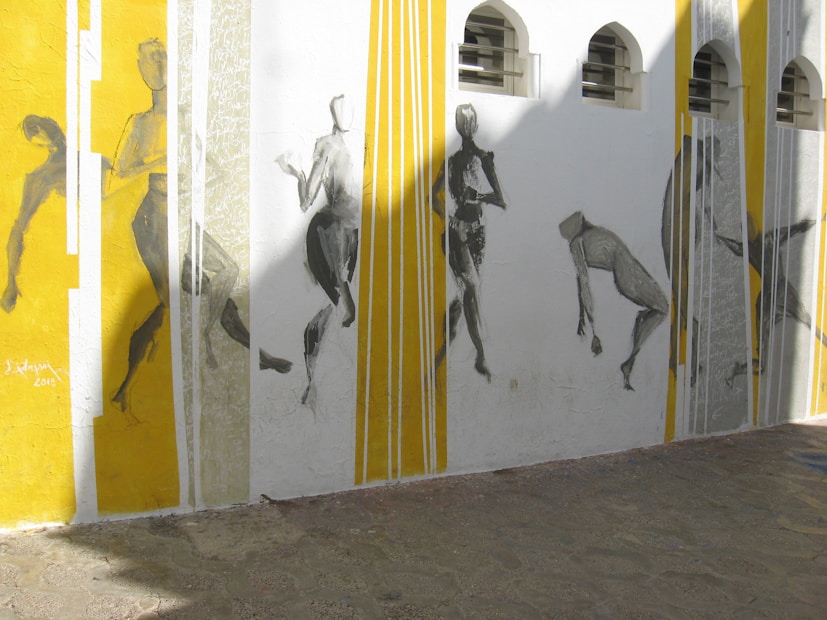 A mural featuring abstract figures painted in monochromatic tones. The figures are in various dynamic poses along a wall segmented by vertical lines in yellow and white. Sunlight casts shadows across the mural, creating an interplay of light and shadow.