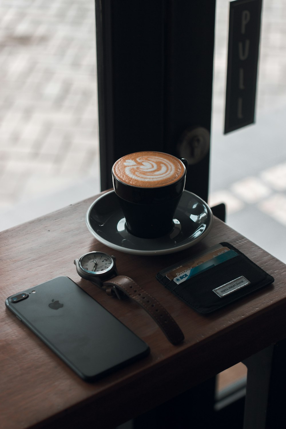 schwarze Keramik-Teetasse mit Kaffee-Latte auf Untertasse neben schwarzem iPhone 7 Plus auf braunem Holztisch