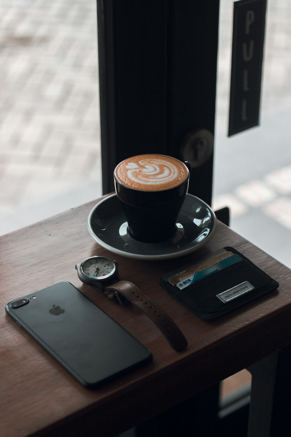 tazza da tè in ceramica nera con latte al caffè sul piattino accanto a iPhone 7 Plus nero su tavolo in legno marrone