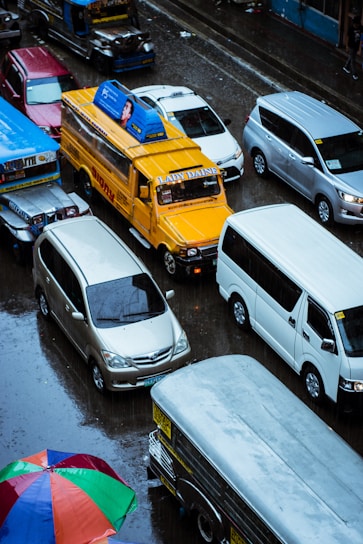 A busy urban street scene features various vehicles, including a distinct yellow jeepney with advertisements on the roof, surrounded by other cars and a colorful umbrella in the foreground. The road appears wet, indicating recent rainfall.