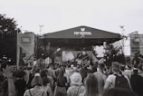 A large crowd of people is gathered in front of a stage at an outdoor music festival. The stage is set up with a canopy labeled 'FSP Festival', and there appears to be a band performing. The atmosphere is lively as the audience engages with the performance. Banners and equipment are set up around the stage, and trees are visible in the background. The image is in black and white.