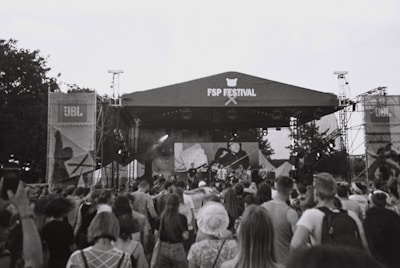 A large crowd of people is gathered in front of a stage at an outdoor music festival. The stage is set up with a canopy labeled 'FSP Festival', and there appears to be a band performing. The atmosphere is lively as the audience engages with the performance. Banners and equipment are set up around the stage, and trees are visible in the background. The image is in black and white.