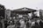 A large crowd of people is gathered in front of a stage at an outdoor music festival. The stage is set up with a canopy labeled 'FSP Festival', and there appears to be a band performing. The atmosphere is lively as the audience engages with the performance. Banners and equipment are set up around the stage, and trees are visible in the background. The image is in black and white.