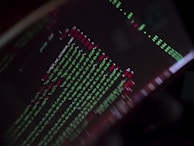 A close-up view of a computer screen displaying programming code with lines of text in green, red, and white against a black background. The code appears to be slightly blurred, creating a moody and abstract visual effect.