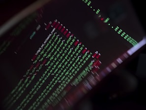 A close-up view of a computer screen displaying programming code with lines of text in green, red, and white against a black background. The code appears to be slightly blurred, creating a moody and abstract visual effect.