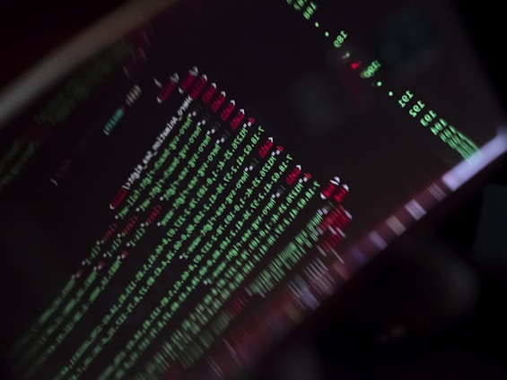 A close-up view of a computer screen displaying programming code with lines of text in green, red, and white against a black background. The code appears to be slightly blurred, creating a moody and abstract visual effect.