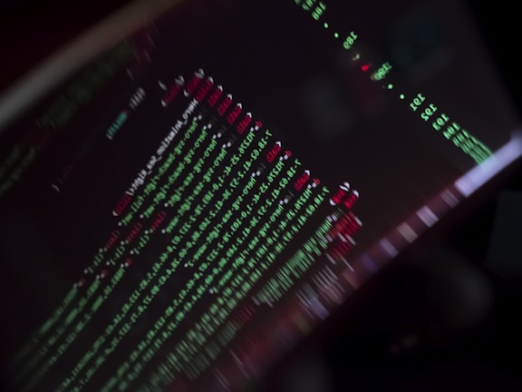 A close-up view of a computer screen displaying programming code with lines of text in green, red, and white against a black background. The code appears to be slightly blurred, creating a moody and abstract visual effect.